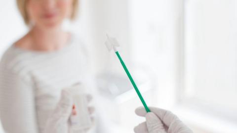 A doctor or nurse's hand can be seen holding cervical screening equipment, while an out of focus woman sits nearby