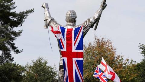 A tall statue with a large union jack attached to its back. There are several smaller union jacks and St George's crosses around it.