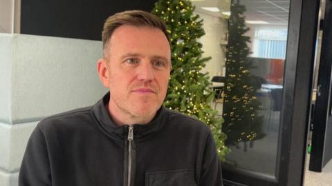 A man with short brown hair and black zip-top with black framed interior doors behind him and a green christmas tree with lights on