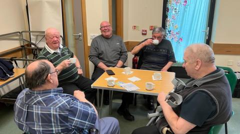 Five men sat around a table in a small meeting room. They all have tea or coffee cups in their hands or on the table.