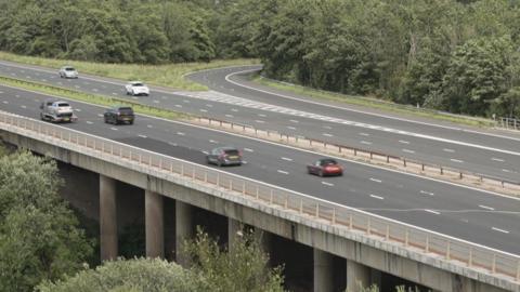 The M6 motorway crossing Castle Howe Bridge in Cumbria. Each carriageway has three lanes and there is a road turning into the motorway on the far side. Many trees line both sides and the circular concrete pillars which hold up the bridge can be seen on the near side.