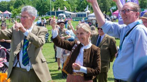 The Duchess of Edinburgh is standing in a grassy area giving a thumbs up flanked by two men doing the same. She is smiling and wearing a brown jacket over a red patterned skirt and has her blonde hair tied back. Behind her are people smiling.