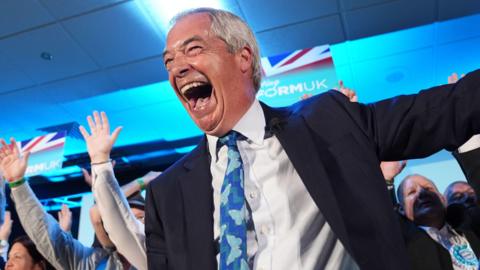 Nigel Farage - a man with silver hair and dressed in a suit with blue tie - laughs happily onstage while surrounded by supporters