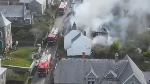 Fire trucks on a road next to a building which is on fire. There is smoke billowing from the roof of the building. There are houses lining either side of the street.