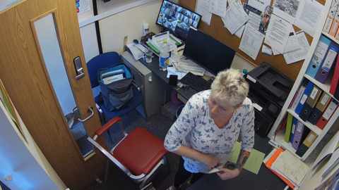 A CCTV image of Jamiel Slaney-Summers. She is wearing a white top with butterflies on. She has short, grey hair and a sleeve tattoo on her left arm. She is in an office. We can see a computer and a chair and paperwork. A door with a lock on is open. She is pulling something out of an envelope. 
