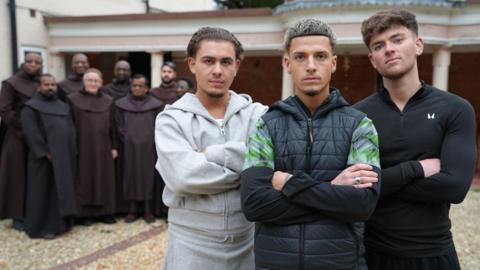 Three young men standing with their arms crossed with a group of traditional friars standing behind them in a courtyard.