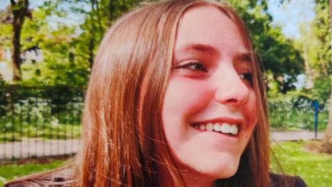 Chloe Longster smiling in a park. You can see her head and shoulders. She is in her school uniform and has long, light brown hair. She is looking to her left.