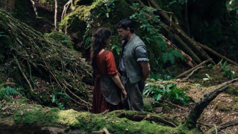 A man and woman standing facing each other in a forest. A woman has long brown hair and is wearing a red dress and the man has short dark hair and a green/ blue shirt.