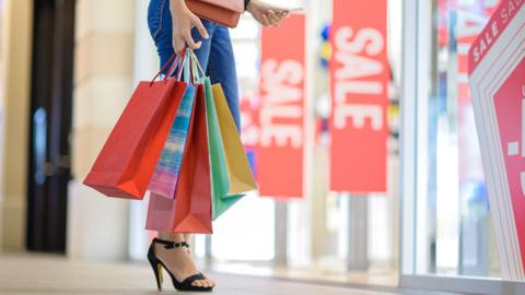A woman wearing jeans and black heels walking through a shopping centre, holding several colorful shopping bags, with large red sale signs displayed in store windows.