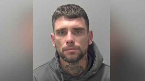 Nathan Staples' police custody picture. He stares expressionless at the camera. He has dark, short hair and wears a grey hoodie. He has a neck tattoo of a triangle with an eye on it. 