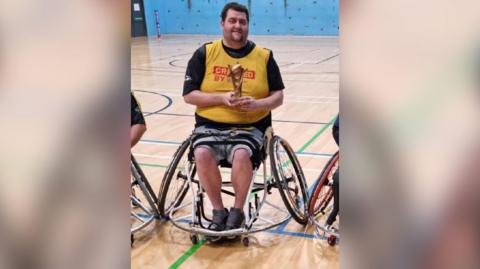 Patrick Murphy, sat in his wheelchair and holding a rugby league trophy in a gymnasium.