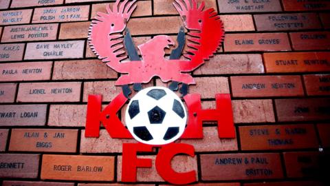 Badge of Kidderminster Harriers with a bird on top of a black and white football with the letters K and H either side of it and FC underneath. The badge is attached to a wall of bricks showing names of supporters.