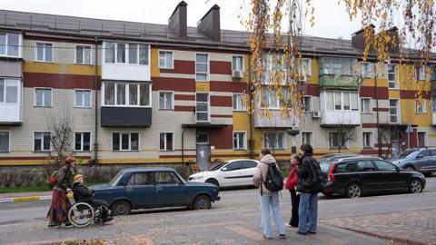 Cars parked on a road, a person being pushed in a wheelchair and a group of women with backpacks standing about, in Bucha