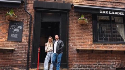 A traditional brick pub exterior with the name ‘The Laddie’ displayed outside. In the doorway there is a man and woman standing close together.  Hanging baskets are placed either side of the doorway and a black sign to the left reads ‘Welcome to the Highland Laddie’.