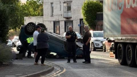 A small hatchback is pictured on its roof as police officers and members of the public stand in front of the vehicle. A lorry and several other cars drive around the wreckage.