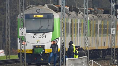 A green-and-white train stopped on a railway track, with officials standing beside it. Overhead power lines and trackside signs are visible in the scene.