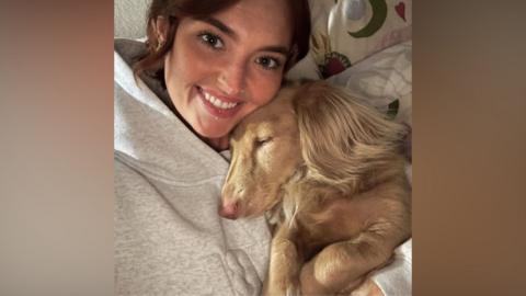 Jess McKee wearing a grey tracksuit cuddles a sandy-coloured dog. She is smiling.