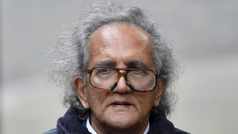 Headshot of Aravindan Balakrishnan arriving at Southwark Crown Court in London in 2015 - mid length grey/black hair balding in the middle, elder man wearing big tortoiseshell glasses, furrowed brow not smiling, wearing a navy coat and a white collar shirt