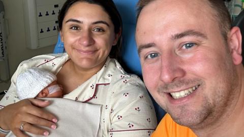 Aisha McCracken in pyjamas in hospital holding her newborn baby who is wearing a white woolly hat and is covered by a blanket. Husband David, wearing an orange T-shirt, is also in the picture. The couple are both smiling at the camera.