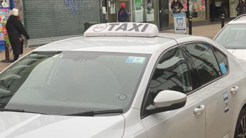 Two white taxis are shown parked in a straight line, with shops on a high street in the background, with people walking in both directions.