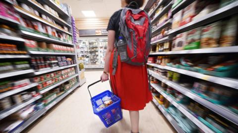 A woman walks down a food shopping aisle holding a basket. The shopper is wearing a red skirt and backpack. The shopping basket is blue.