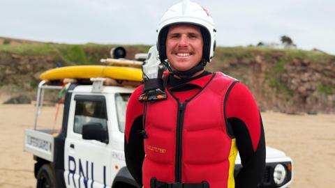 Will Simmons is smiling and standing in front of a RNLI-branded truck. He has a red life jacket on and is wearing a white helmet.