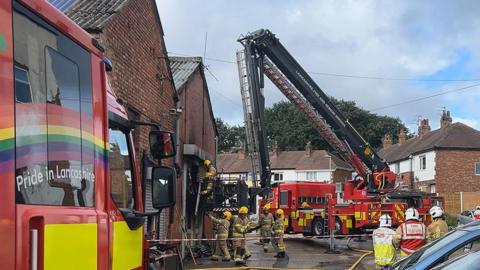 Firefighters inside a cordon tackling a fire at a storage unit on an industrial estate with two fire engines one with an aerial platform close by and a row of houses nearby.