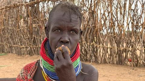 A woman with beaded, colourful necklaces around her neck is putting some food in her mouth.