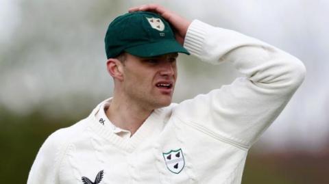 Worcestershire bowler Ben Gibbon puts his left hand on top of his green club cap as he looks at the action while in the field