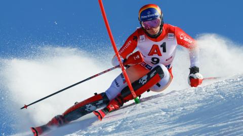 Alpine skier in red and white racing suit carving through snow on a steep slope, leaning sharply with poles extended, snow spraying behind against a clear blue sky.