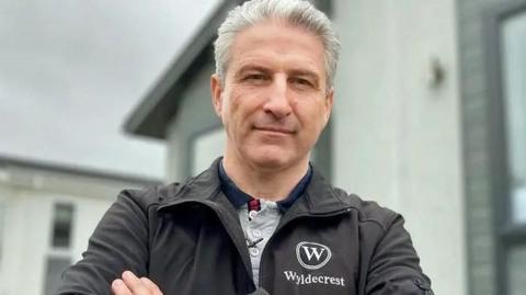 A middle aged man with grey swept back hair stands with his arms crossed wearing a black jacket with a logo which reads Wyldecrest. Underneath it he is wearing a grey polo shirt. A mobile home is in the background. 