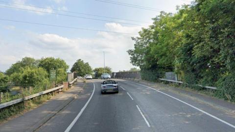 Cars travel over a railway on a road which is bordered by trees either side. There is a footpath over the railway and an opening to some steps on the left hand side.