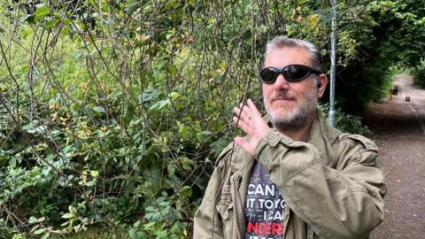 A visually impaired man, wearing dark sunglasses, an olive jacket and a navy t-shirt, puts his hand up to feel long thin branches that have grown on to the path