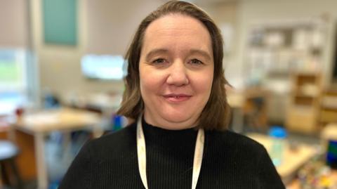 Steph Smith looking into camera in a classroom. She has shoulder-length brown hair and is wearing a black jumper with a white lanyard.