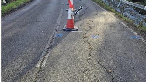 A tarmac road with large cracks with traffic cones along it. Some fencing is falling off the side of the road.