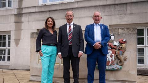 Two men in suits and a woman in garish blue trousers. 