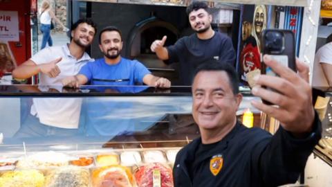 A man holds his phone up to take a selfie of himself and three men. The men stand behind a food counter with their thumbs up. The man taking the picture wears a Hull City jacket and smiles at the camera.