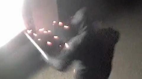 Still from CCTV showing a man in a black coat with his hood up holding a stick giving off sparks next to a cash machine