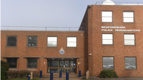 Front of Bedfordshire Police headquarters. A red-brick building with large windows and "Bedfordshire Police Headquarters" written on the front of it. It has a number of car bollards outside its front door. 