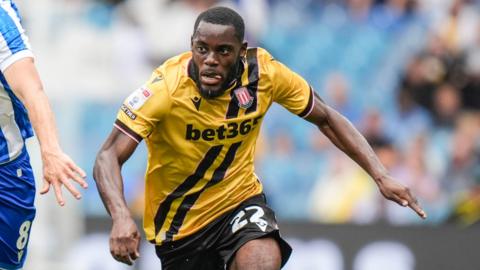 Junior Tchamadeu is mid-run and has a focused expression on his face. His tongue slightly pokes out to the left side of his mouth. he wears a dark yellow shirt with two black diagonal stripes down the middle and black shorts.