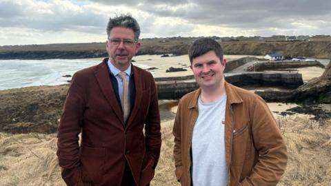 Dithis fhireannach nan seasamh le an làmhan nam pòcaidean. Tha an tràigh air an cùlaibh.