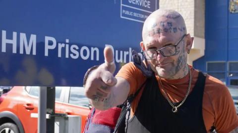 Jason Hoganson standing in front of a sign for HM Prison Durham with his thumb up. He is bald with a tattooed hand, arm, head and face.