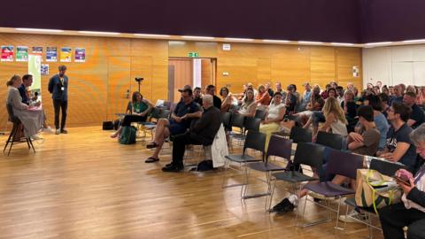 Dozens of people sat inside a school hall for an event. Two people with microphones are sat in front of the audience.