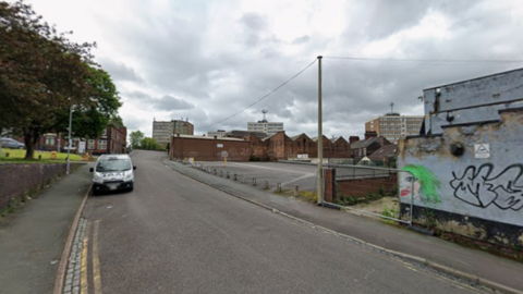 General view of Hillcrest Street in Hanley. An empty street with an empty car park on the right and a dilapidated blue building with graffiti on. On the left parked on the side of the road there is a silver taxi
