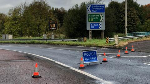 A road junction with orange cones across the lane with a blue sign saying "police accident". There is a 40 mph speed limit sign in the background and a road sign with directions to Londonderry, Cookstown, Ballymena, Coleraine and Belfast.