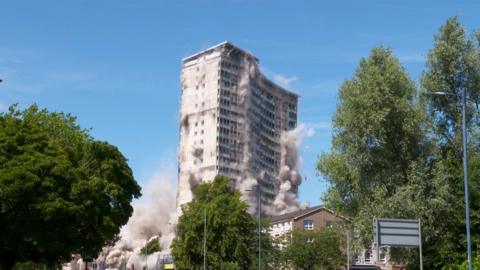 A high-rise tower block mid- demolition. The building is in the middle of collapsing and there are plumes of dust coming out of it. It sits against a blue sky and there are tall trees at either side of the image.