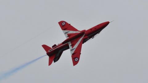 A red jet with white markings flies with blue smoke spraying off its back