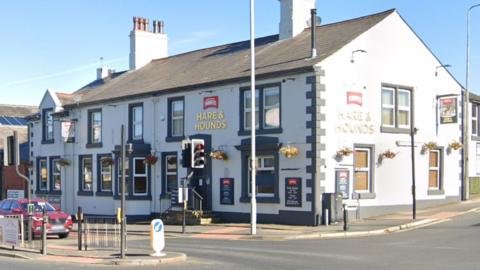 Street view of the Hare and Hounds. A large white rendered two storey pub on the corner of a road junction by traffic lights and dark grey painted window surrounds