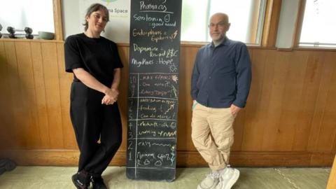 Two people standing either side of a long blackboard headed “The Body Pharmacy” with scores for effort underneath.