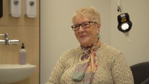 A woman looks slight off camera. She has glasses and short white hair. She is wearing a cream woolly jumper with a colourful scarf. She is sat in what looks like a hospital room.